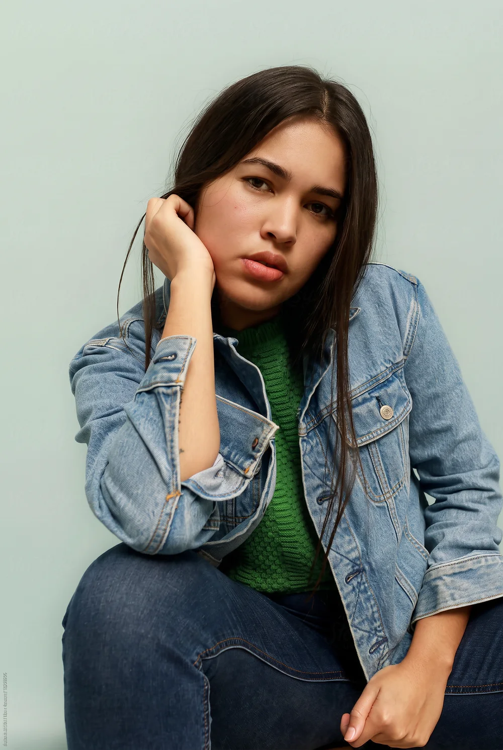 Young adult in a denim jacket and green top against a mint studio background